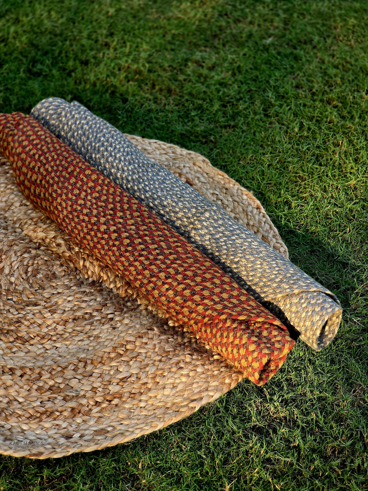 Two braided rugs in orange and brown colors are displayed on a round, beige straw mat, placed on a grassy surface.