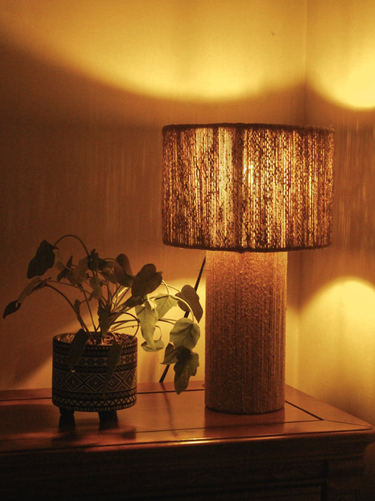 Decorative Jute Table lamp with a textured shade on a wooden surface next to a plant, casting a warm glow.