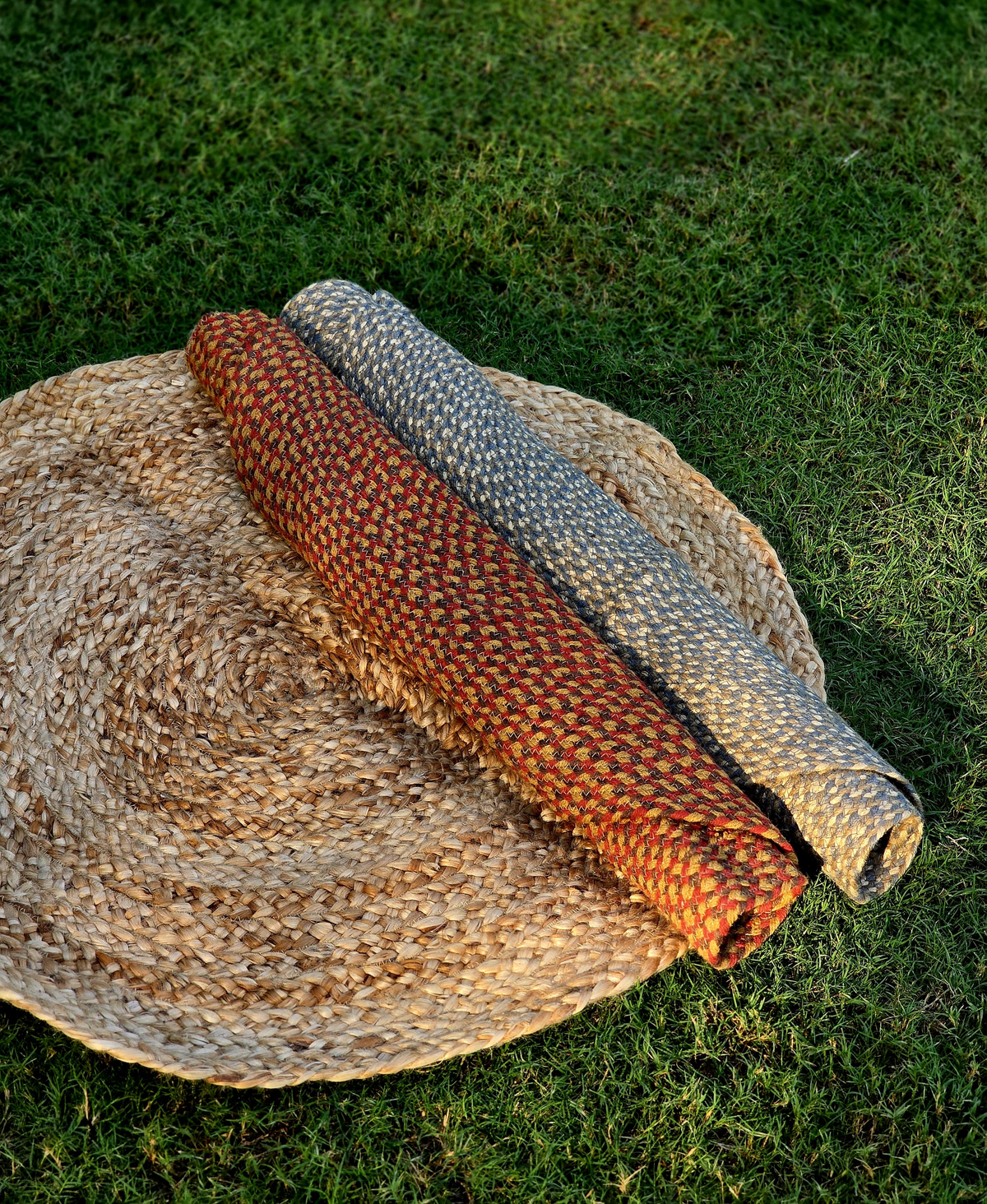Stacked round jute rugs folded and placed on a larger round jute rug on a green park lawn, showcasing natural outdoor eco-friendly styling.