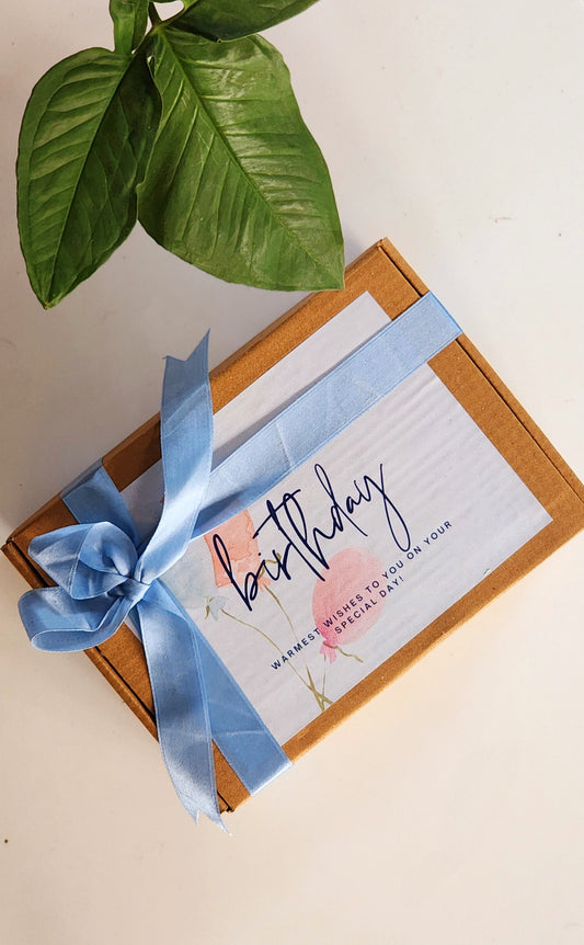 A handmade birthday hamper gift set with a candle, cup, and bowl, tied with a blue ribbon and a 'Happy Birthday' message on the front, presented on a white surface with a green leaf and a coconut shell decoration in the background.