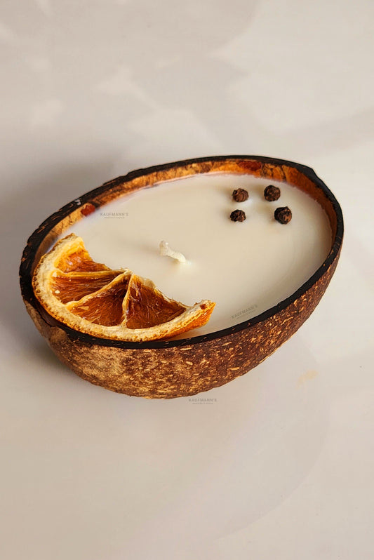 A coconut shell candle with the inner part of the shell exposed, with an orange slice and some dried leaves on top, placed on a white surface.