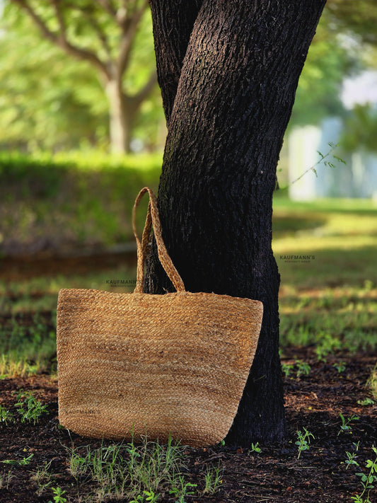 A beige coir tote bag hanging on a tree branch in a natural outdoor setting.