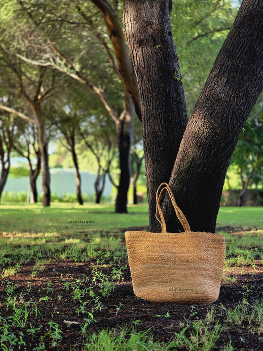 A beige coir tote bag hanging on a tree branch in a natural outdoor setting.
