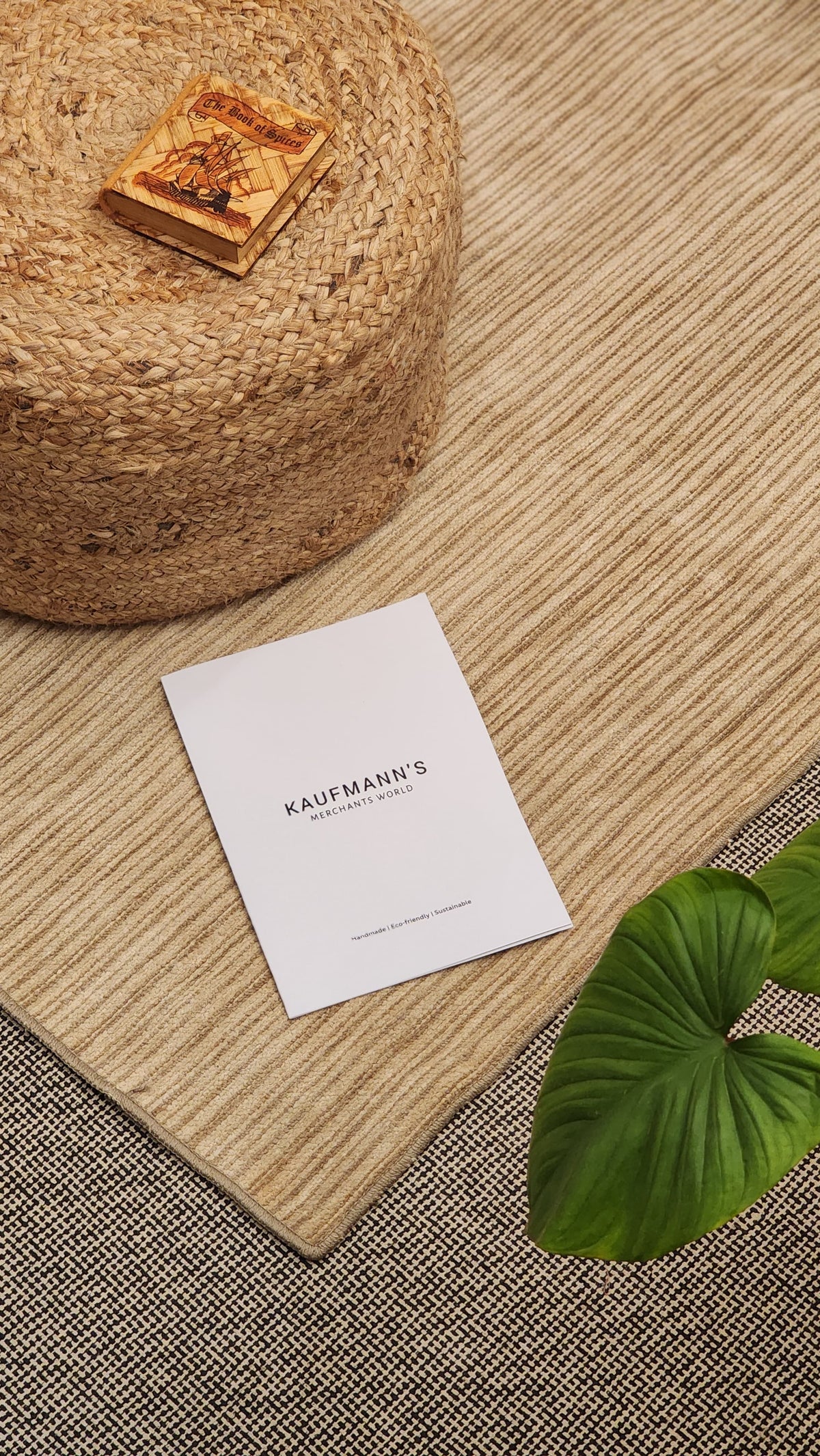 A beige cotton rug with a flatweave pattern, placed on a floor next to a round jute rug, a cardboard box with a design, and a business card with the text 'Kaufmann's'. The background is out of focus.
