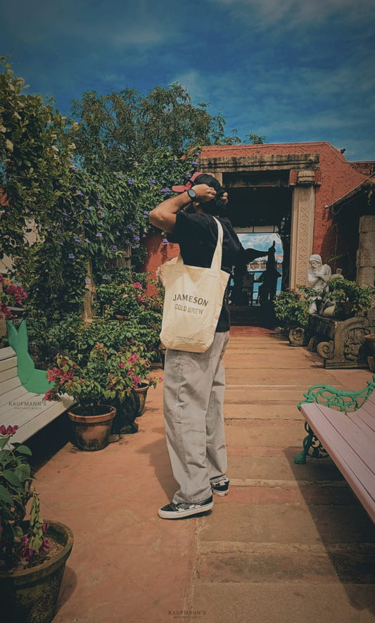 A person standing on a pathway in a garden-like setting, looking back over their shoulder at the camera. They are holding a beige cotton tote bag with a logo visible on the front.