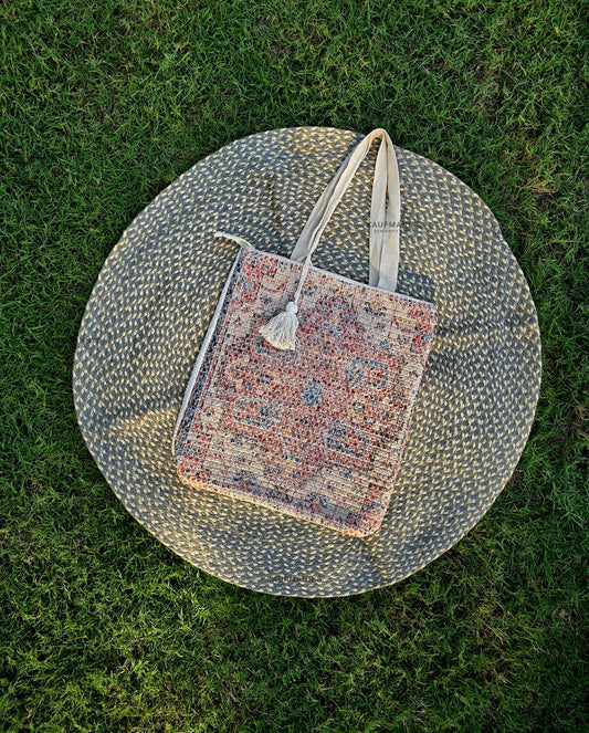 A jute tote bag with multicolor pattern, placed on a round beige textile surface, set against a grassy background.