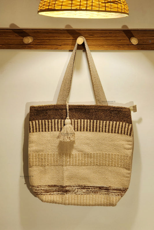 A beige Jute tote bag hanging on a wooden hanger against a white wall, with a handle and a drawstring closure visible.