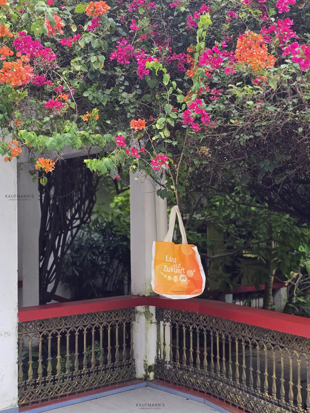 An orange cotton tote bag hanging over a balcony railing, with a lush pink and green floral background.