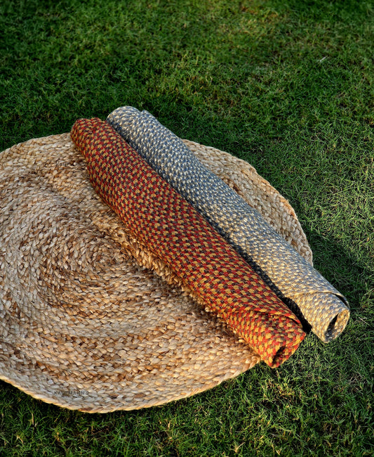 Two braided rugs in orange and brown colours are displayed on a round, beige straw mat, placed on a grassy surface.