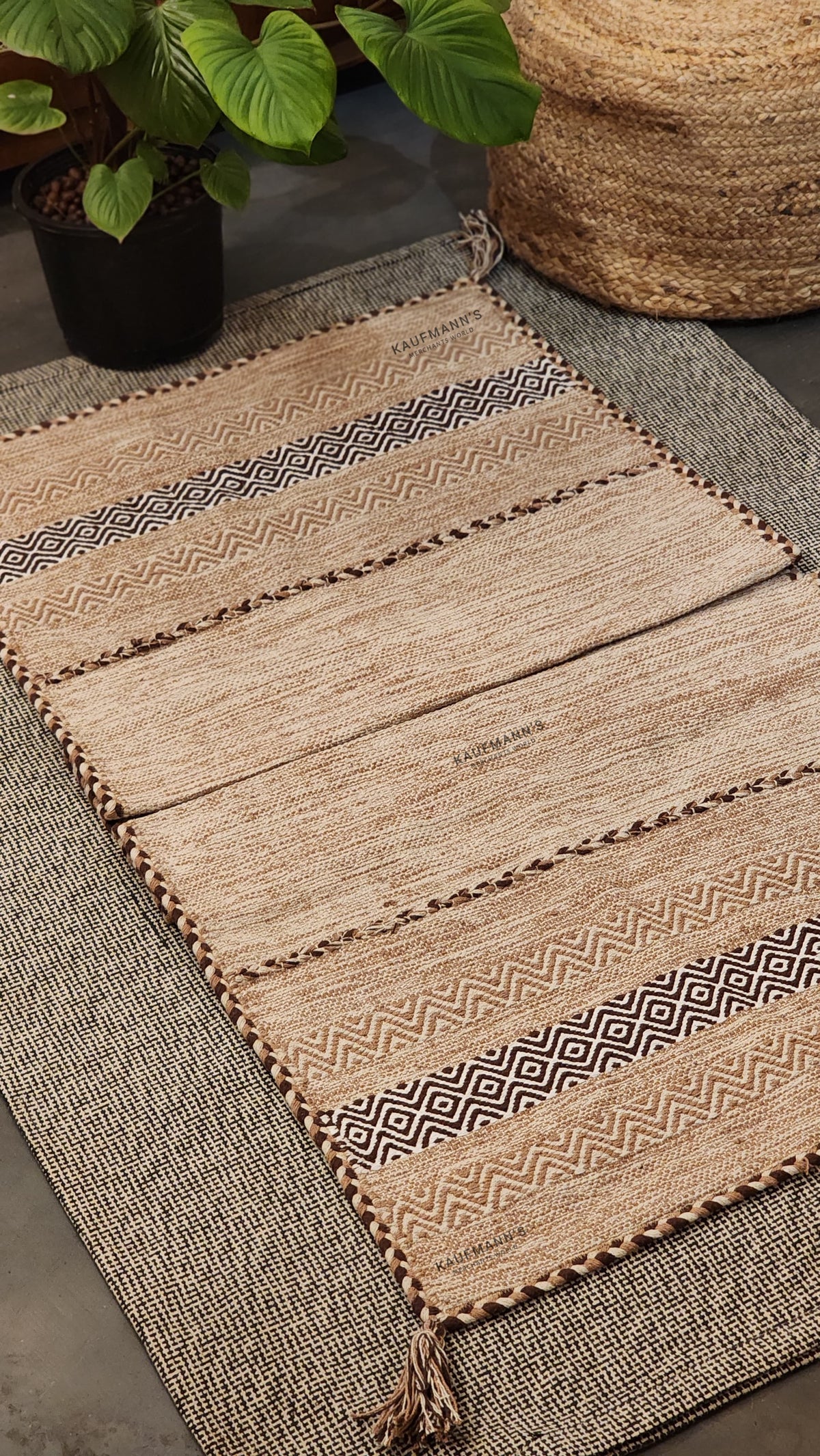 A beige cotton rug with geometric patterns, placed on a floor under a woven mat, with plants and a wicker object in the background.