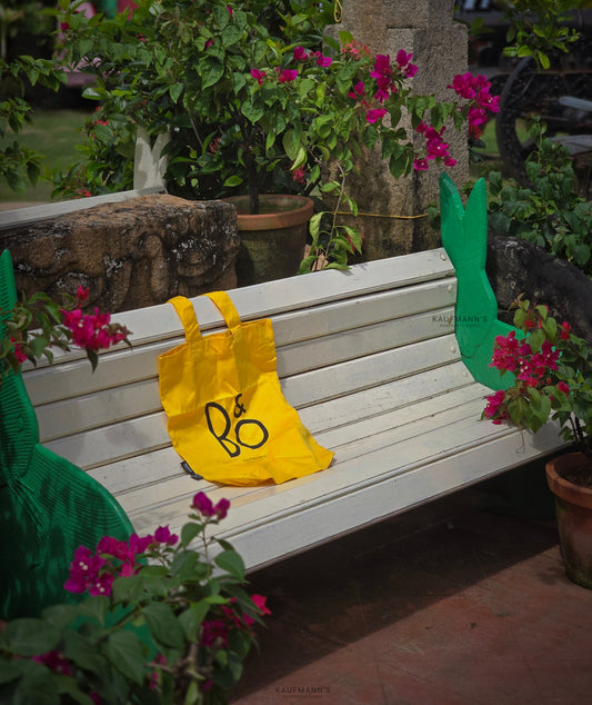 A yellow cotton tote bag with a black logo placed on a white bench in a garden setting, surrounded by potted plants and flowers.