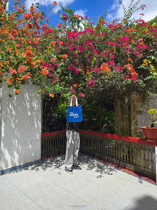 A person standing on a balcony with a vibrant pink and green floral background, holding up a blue cotton tote bag in front of their face.