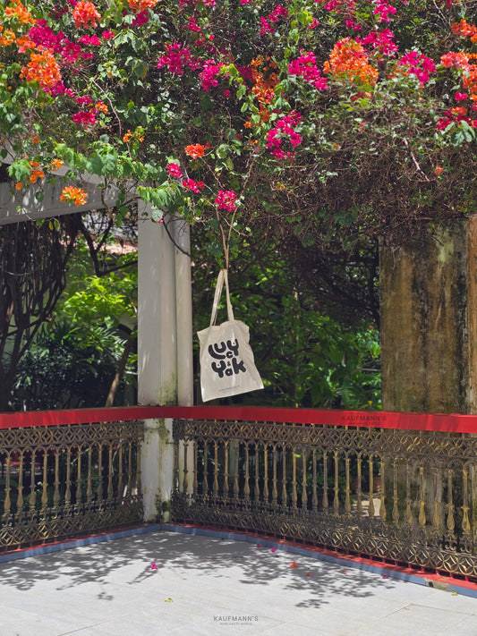 A white cotton tote bag hanging over a red railing, with a vibrant pink floral background.